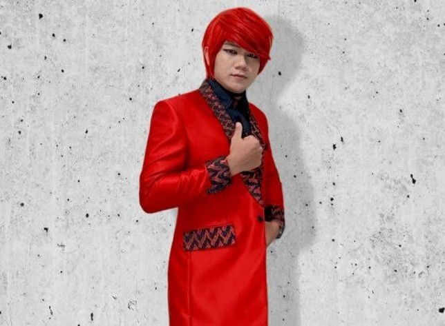 Pesulap Merah berambut merah mengenakan setelan jas merah sambil mengacungkan jempol