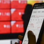Tangan memegang ponsel yang menampilkan grafik saham di depan papan perdagangan berwarna merah