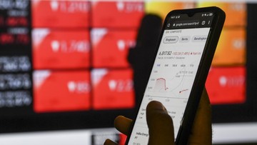 Tangan memegang ponsel yang menampilkan grafik saham di depan papan perdagangan berwarna merah
