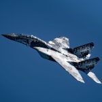 Military MiG-29 jet displaying digital camouflage pattern in clear blue sky.