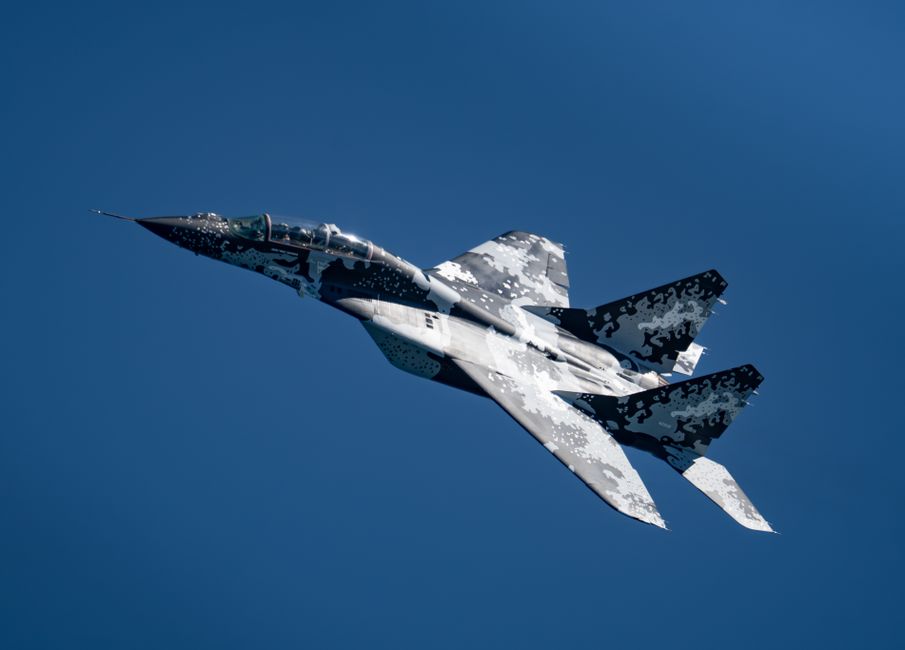 Military MiG-29 jet displaying digital camouflage pattern in clear blue sky.