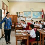 Seorang guru perempuan mengajar di depan papan tulis di ruang kelas dengan murid-murid berseragam putih merah