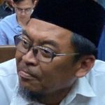 Seorang pria berpeci hitam dan berkacamata duduk di kursi dalam ruangan