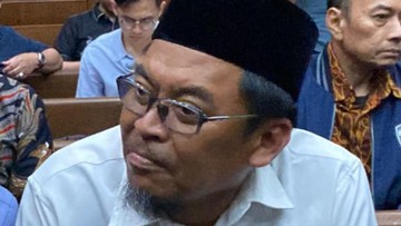 Seorang pria berpeci hitam dan berkacamata duduk di kursi dalam ruangan