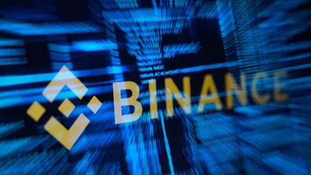 Logo Binance dengan latar belakang teks komputer berwarna biru yang buram