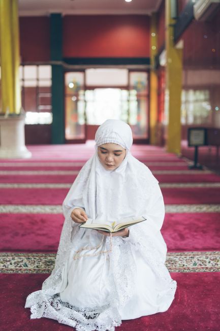 Perempuan muda berhijab putih membaca Al-Qur'an di dalam masjid