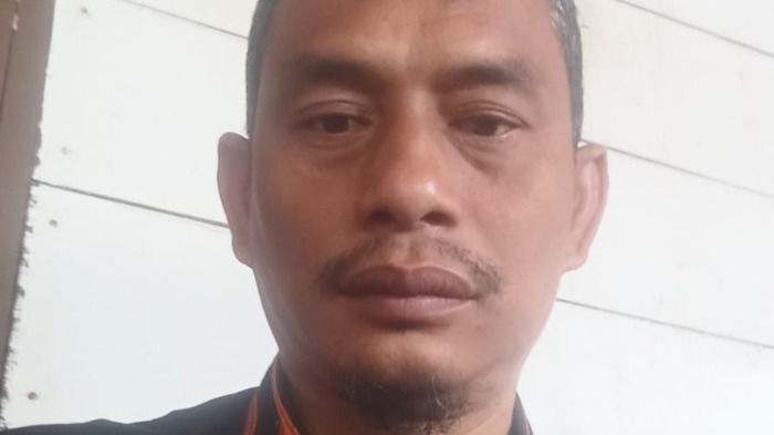 Potret close-up seorang pria berambut pendek dan berkumis memakai baju gelap beraksen oranye