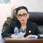 Seorang perempuan berkacamata dan berblazer hitam duduk di meja rapat dengan mikrofon dan botol air