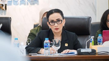Seorang perempuan berkacamata dan berblazer hitam duduk di meja rapat dengan mikrofon dan botol air