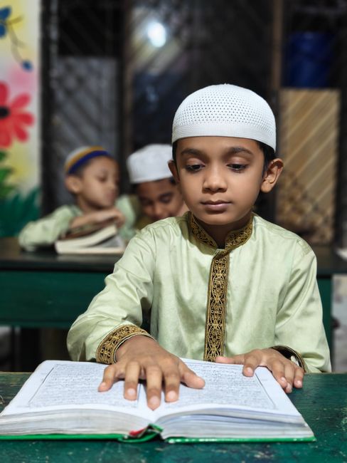 A young boy in a traditional outfit reading the Quran in a classroom setting.