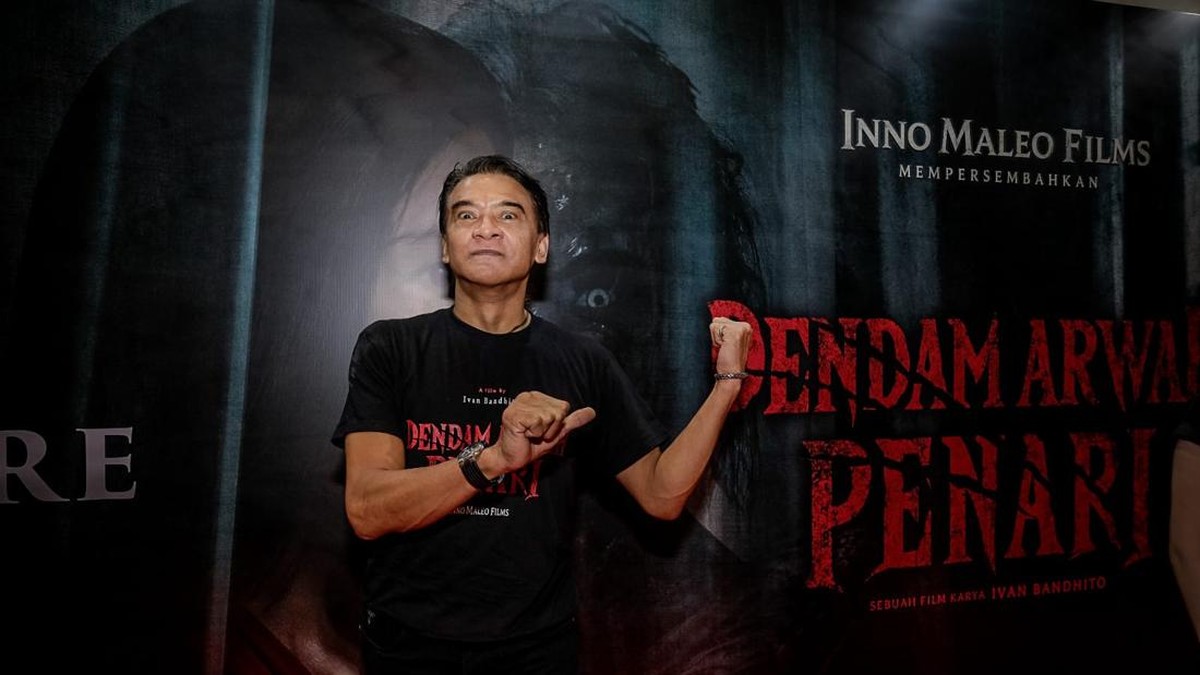 Donny Damara berkaus hitam Dendam Arwah Penari berpose di depan latar film horor Dendam Arwah Penari