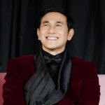 Vino G. Bastian tersenyum memakai setelan merah marun saat duduk di sofa
