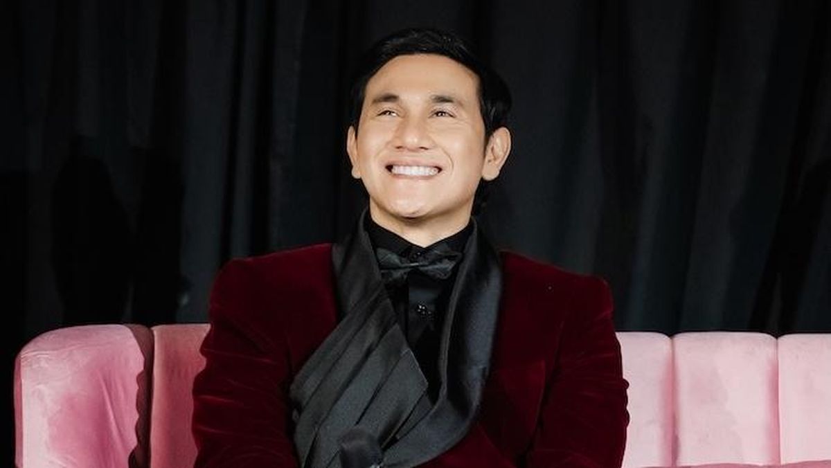 Vino G. Bastian tersenyum memakai setelan merah marun saat duduk di sofa