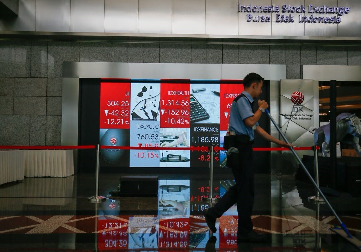 Papan indeks Bursa Efek Indonesia menampilkan angka-angka merah yang menurun dan seorang pekerja membersihkan lantai