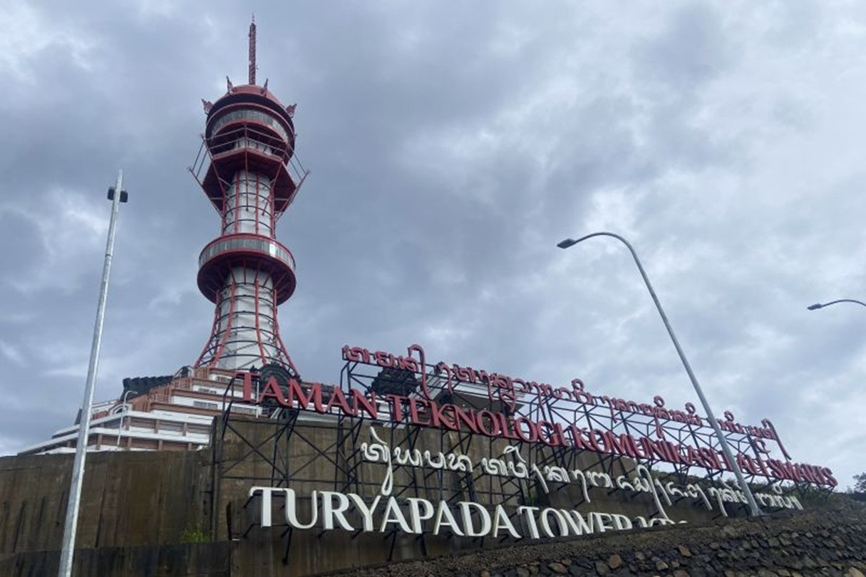 Menara Turyapada, menara komunikasi merah-putih bertingkat dengan dek observasi, difoto dari bawah dengan papan nama Turyapada Tower di depannya