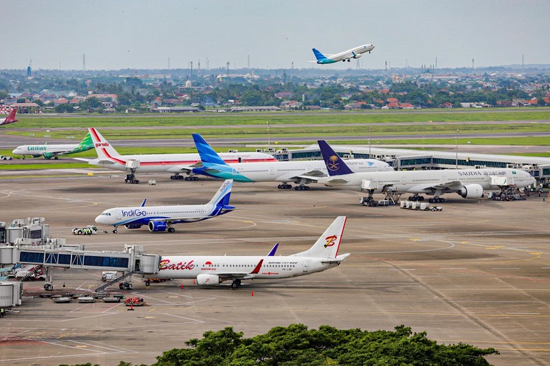 Sejumlah pesawat maskapai terparkir di apron bandara, dengan satu pesawat sedang lepas landas di latar belakang