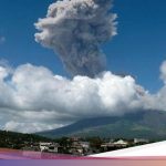 Kolom abu vulkanik pekat membumbung tinggi dari Gunung Mayon di Filipina dengan latar langit biru dan permukiman di kaki gunung
