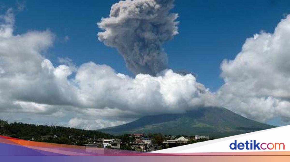 Kolom abu vulkanik pekat membumbung tinggi dari Gunung Mayon di Filipina dengan latar langit biru dan permukiman di kaki gunung