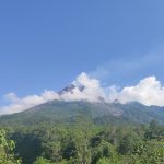 Puncak Gunung Merapi yang diselimuti awan di atas lereng hijau pada hari cerah