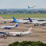 Sejumlah pesawat maskapai terparkir di apron bandara, dengan satu pesawat sedang lepas landas di latar belakang