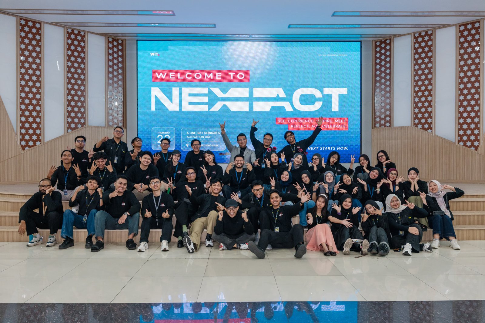 Sekelompok besar karyawan WIT.ID berfoto bersama di depan layar bertuliskan Welcome to Nexact pada acara ulang tahun ke-16