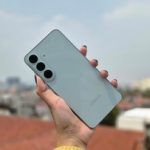 Tangan memegang ponsel Samsung Galaxy berwarna terang dengan tiga kamera belakang di atas latar kota