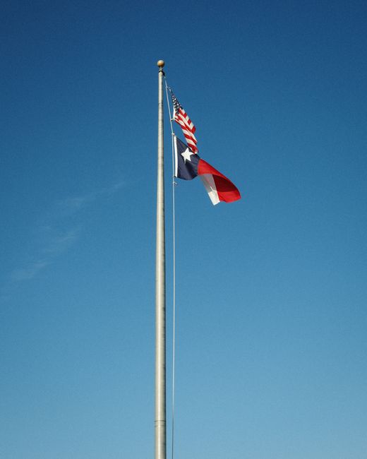 Bendera Texas dan bendera Amerika Serikat berkibar pada tiang tinggi di bawah langit biru cerah